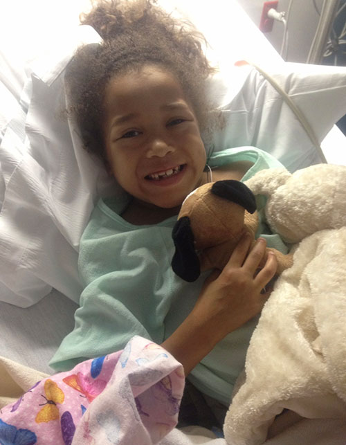 Young girl holding stuffed dog while in hospital bed.