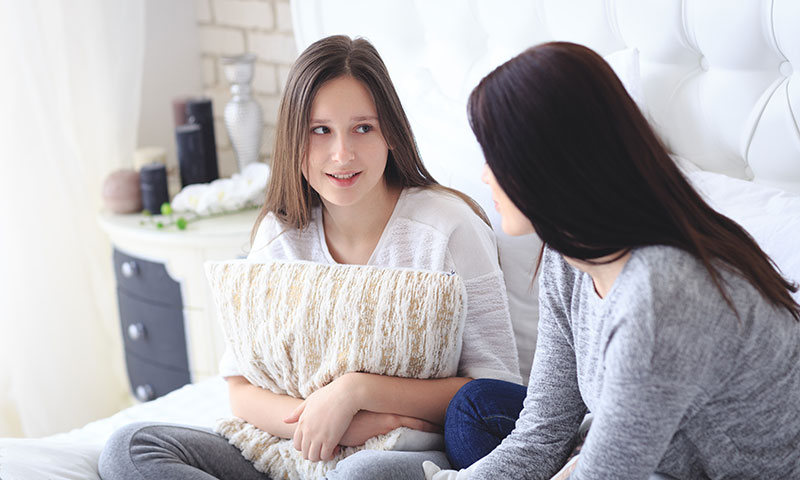 Mother talking with teenage daughter sitting in bed while daughter holds pillow