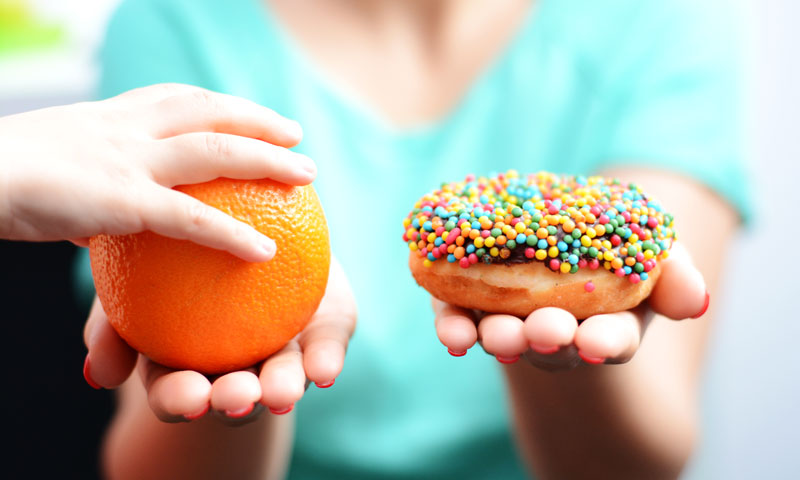 Mother holding an orange in one hand and a donut in the other, child's hand reaches to pick the orange